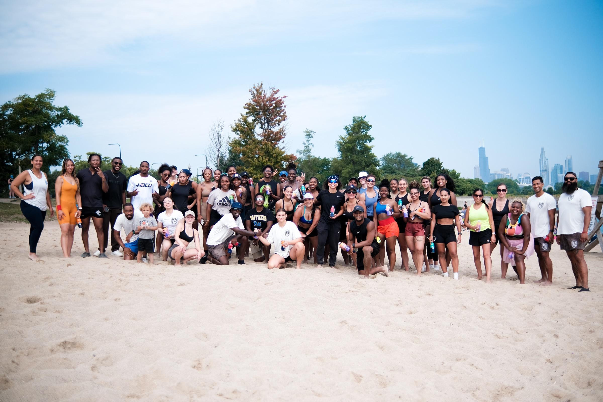 A large group of Body by MJ members and coaches posing together on the beach with the Chicago skyline behind them
