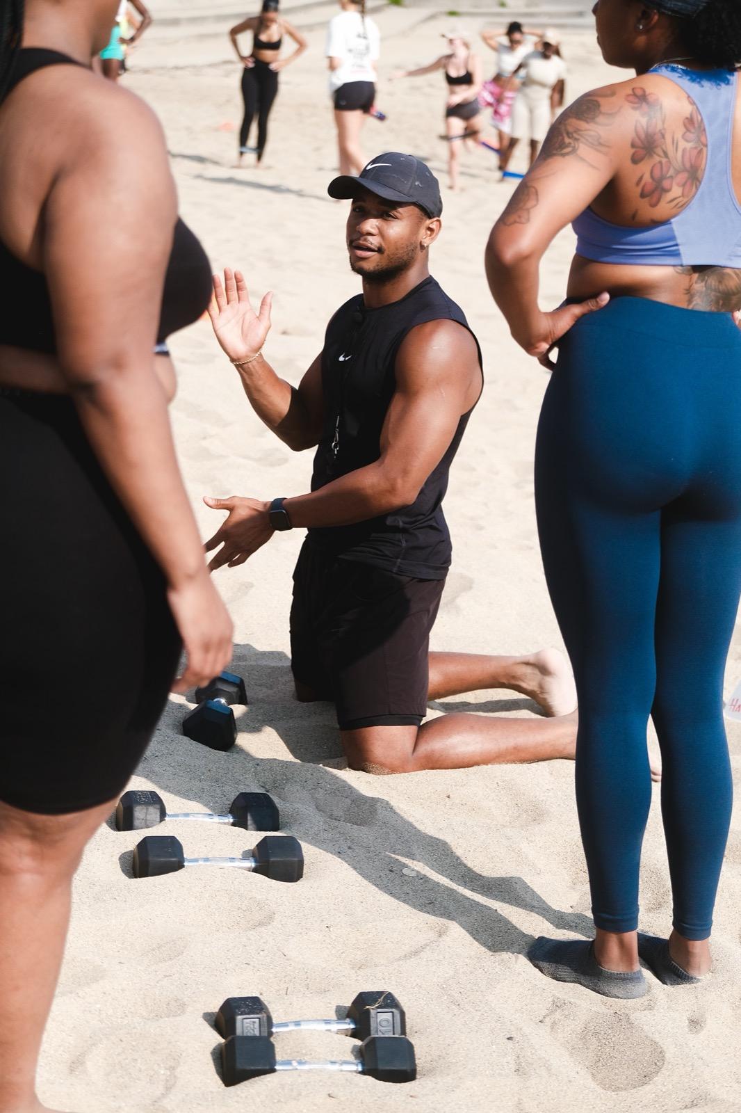Marcus Jones kneeling and coaching members through a dumbbell exercise during a beach class