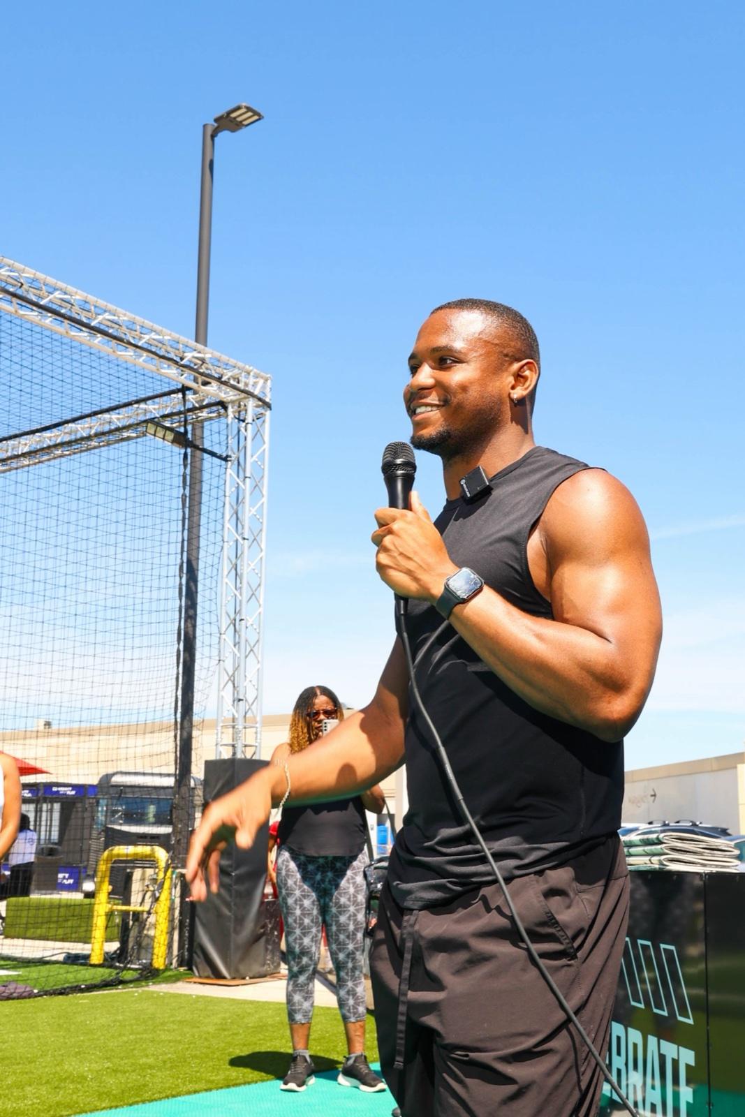 Marcus Jones speaking into a microphone at a Body by MJ event, mid-sentence and smiling